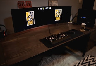 A gaming setup featuring a large curved monitor displaying PUBG logos on the screens. A keyboard and mouse are positioned on a large desk pad. A pair of headphones rests on the desk alongside a mobile phone. A drink in a Starbucks cup is placed to the right, while a desktop tower is seen in the background on the right side.