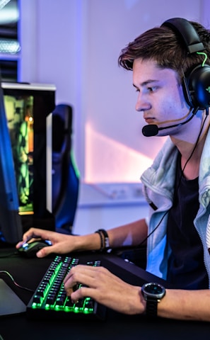 A young person is intensely focused on a computer screen while wearing a headset with a microphone. They are using a gaming keyboard and mouse, sitting in a gaming chair. The room has cool lighting, with a visible PC tower.